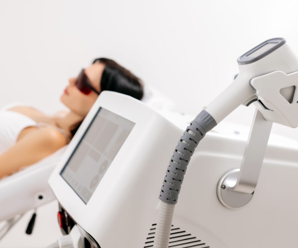 Body care concept. Laser hair removal. Young Caucasian woman in safety glasses lies on the sofa, female cosmetologist doing laser hair removal in a beauty salon